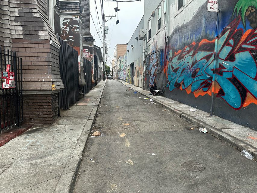 Narrow urban alley with graffiti on the walls, litter scattered on the ground, and buildings lining both sides under overcast skies.