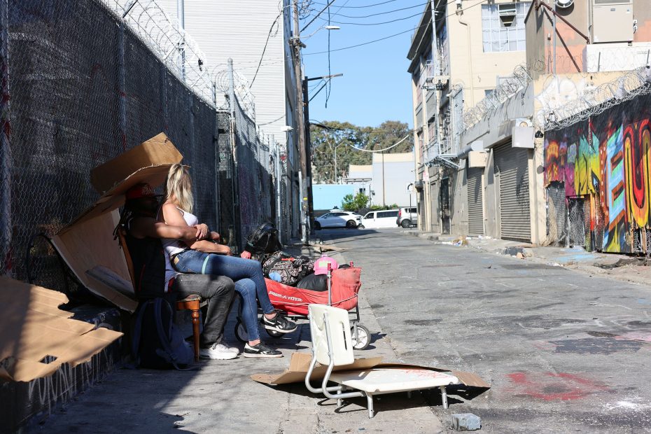 Two people sit on a sidewalk beside a chain-link fence and makeshift shelter in an urban alley with graffiti and debris scattered around.