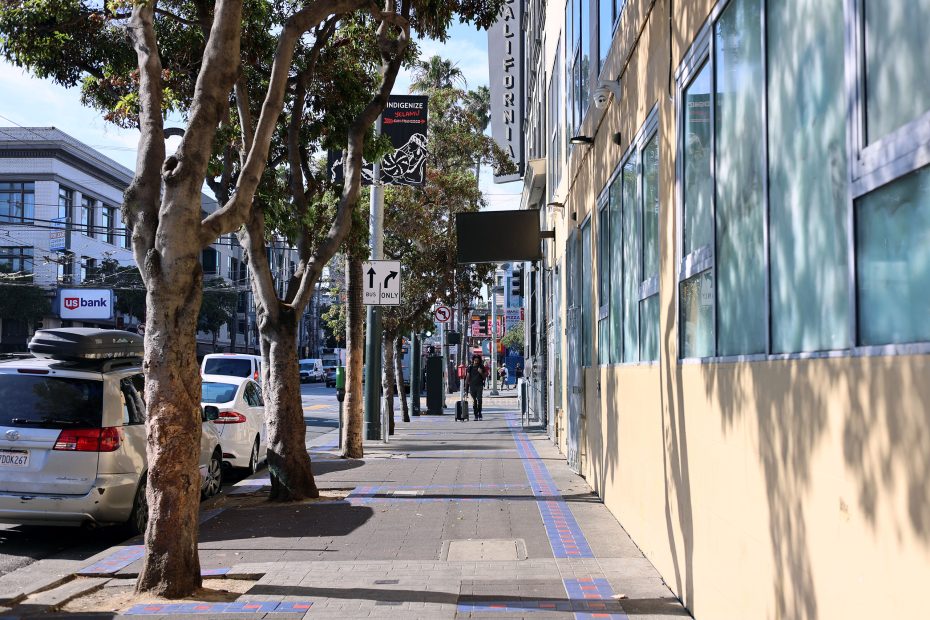 A city sidewalk lined with trees and parked cars runs alongside buildings; street signs and banners are visible, with a few people walking in the distance.