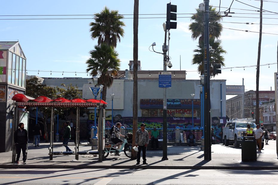 People walk and stand near a street corner with palm trees, outdoor exercise equipment, and colorful graffiti on the building in the background.