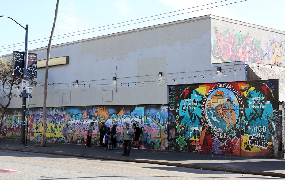 A city street with people walking past a building covered in colorful graffiti and a large mural reading "Indigenous 365" and "Welcome to Tongva Land.