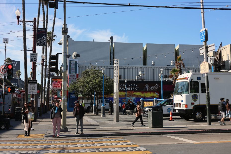 People wait at a crosswalk in an urban area with a police vehicle parked nearby and a mural reading “American Indian Cultural District” in the background.