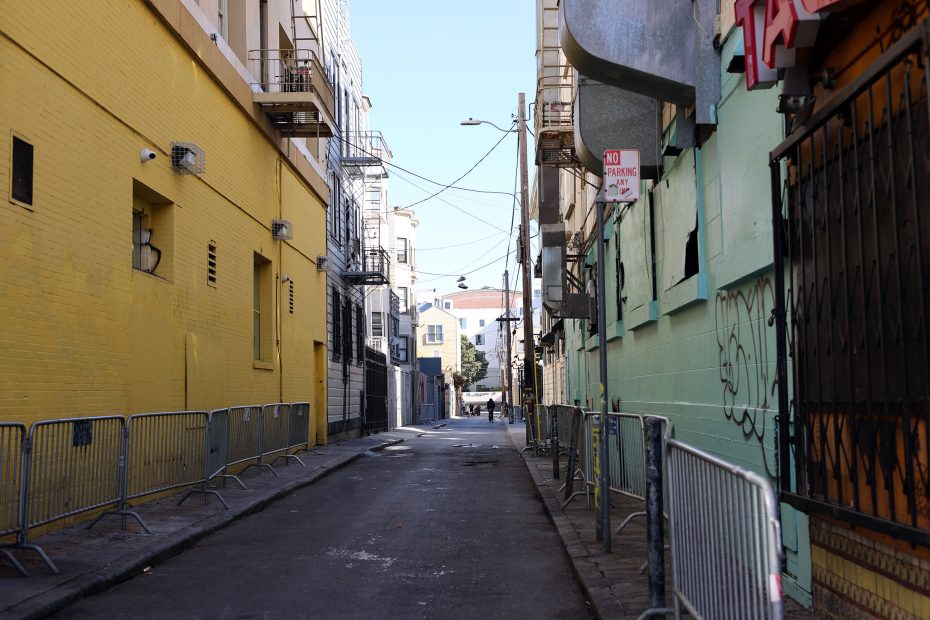 A narrow urban alleyway lined with metal barricades, yellow and green painted buildings, and a no parking sign, with apartments and clear sky in the background.