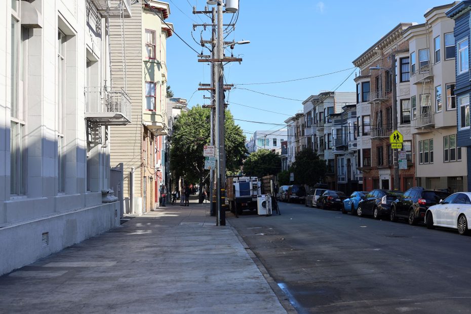 A city street lined with parked cars and multi-story apartment buildings, with a utility truck parked near the sidewalk and people walking in the distance.
