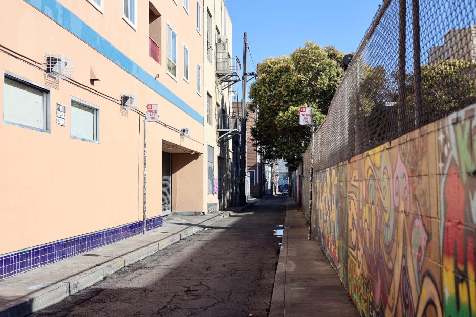 Narrow urban alleyway with a mural-covered fence on one side and an apartment building on the other, under clear blue sky.