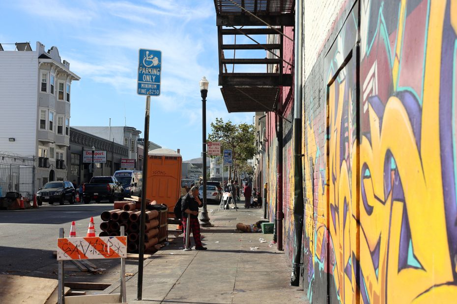 A city sidewalk with graffiti on a building, stacked pipes, a parking sign, and people walking along the street on a sunny day.
