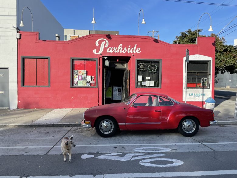 A red vintage car is parked in front of a red building labeled "Parkside." A small dog stands on the street in the foreground, and posters are visible in the building’s window.