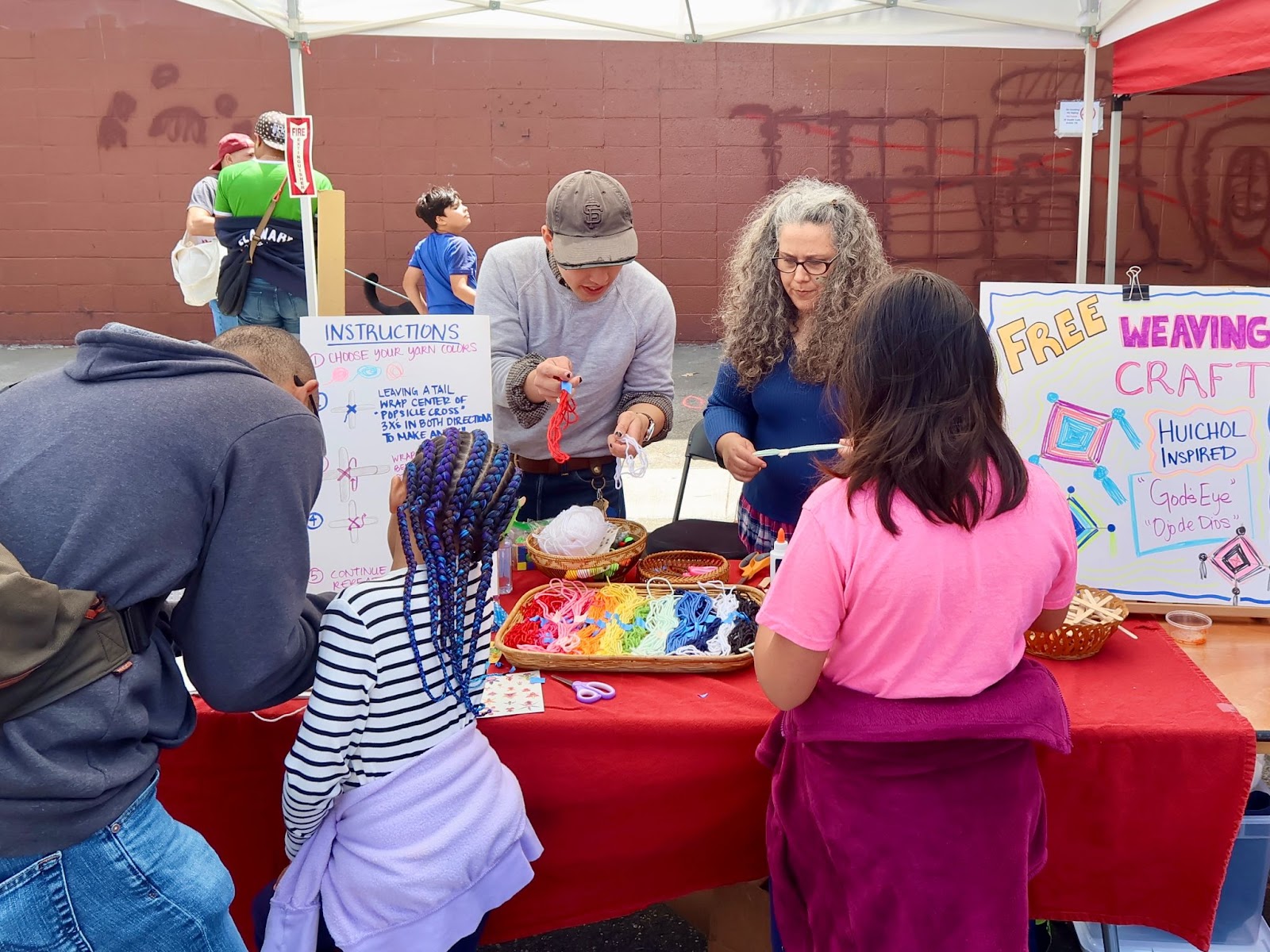 Adults and children participate in a weaving craft activity at an outdoor booth with colorful yarn and instructional signs on display.