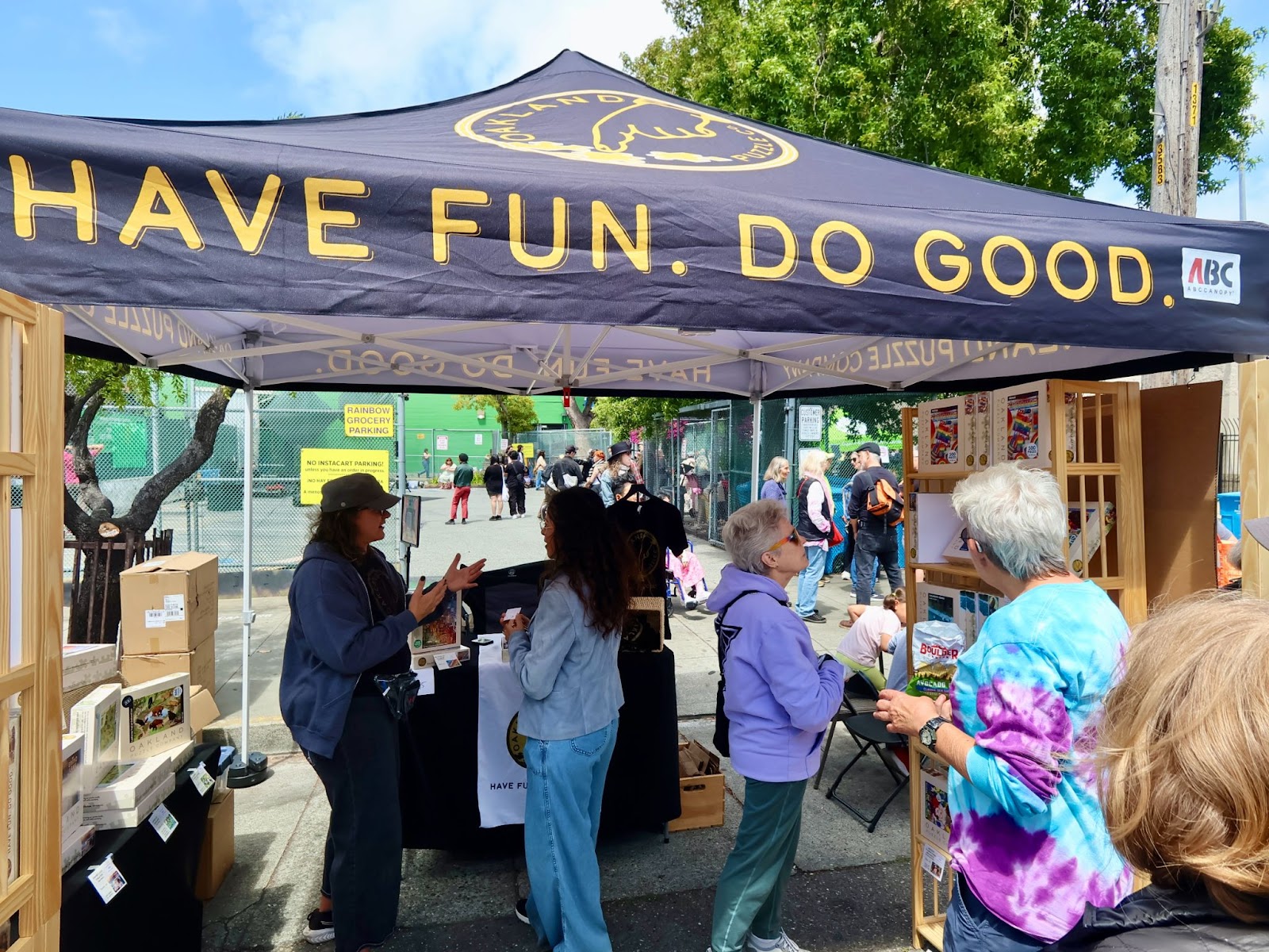 People browsing and interacting at an outdoor booth under a tent with the words "HAVE FUN. DO GOOD." printed on it, on a sunny day.
