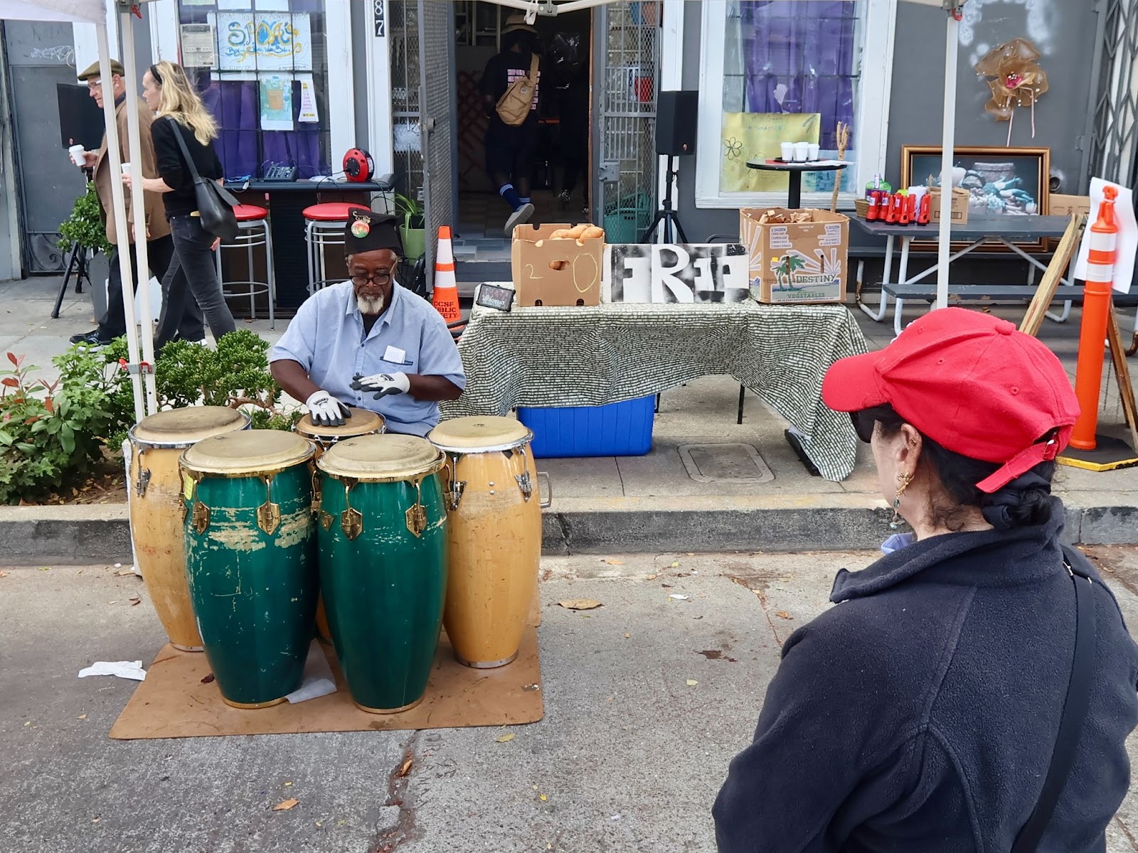 A man plays four conga drums on a sidewalk while a woman in a red cap watches; behind them is a table with a “FREE” sign and various items.