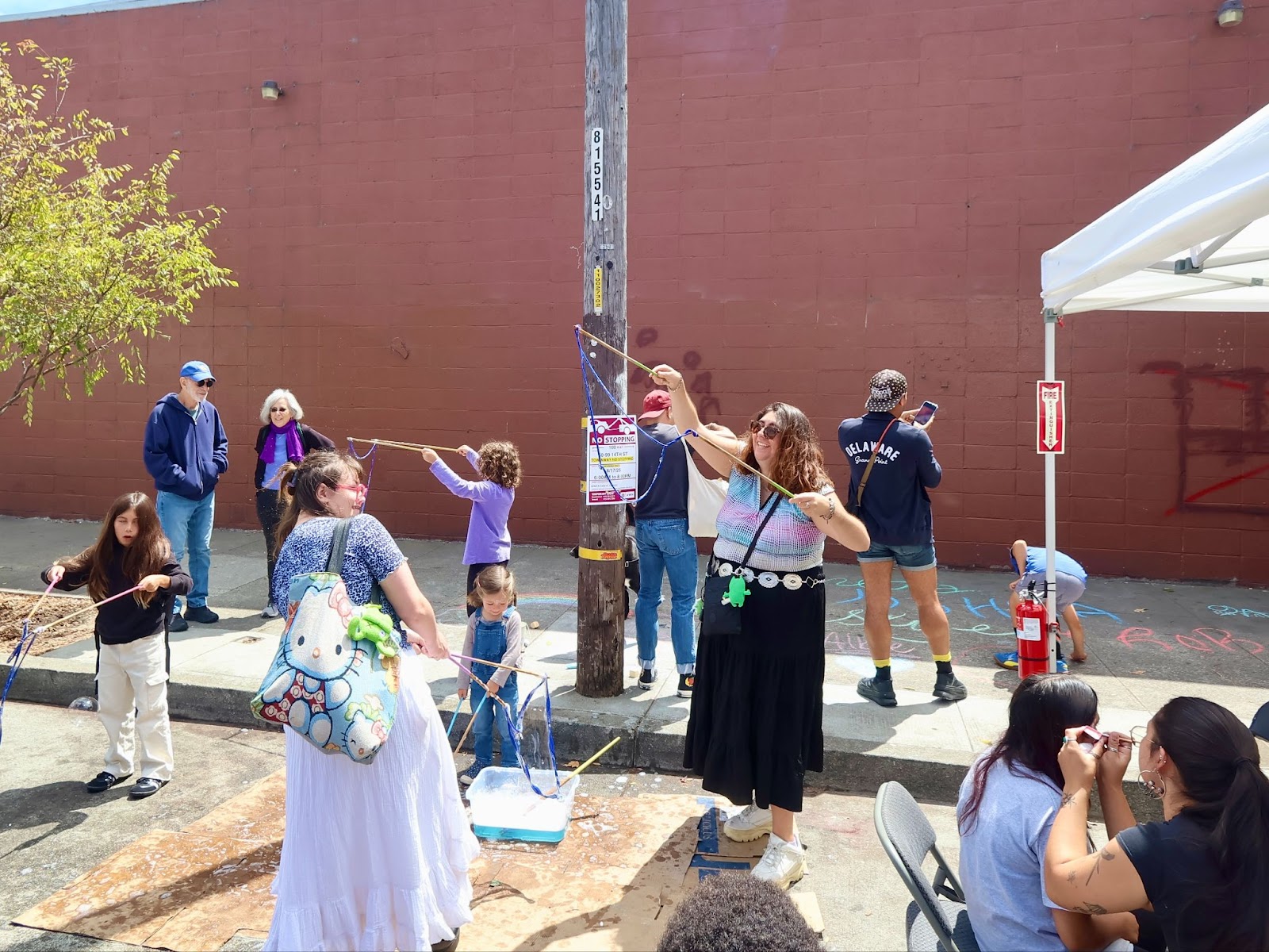 People make large soap bubbles at an outdoor event, with children and adults participating. A tent, chalk drawings, and bystanders are visible in the background.