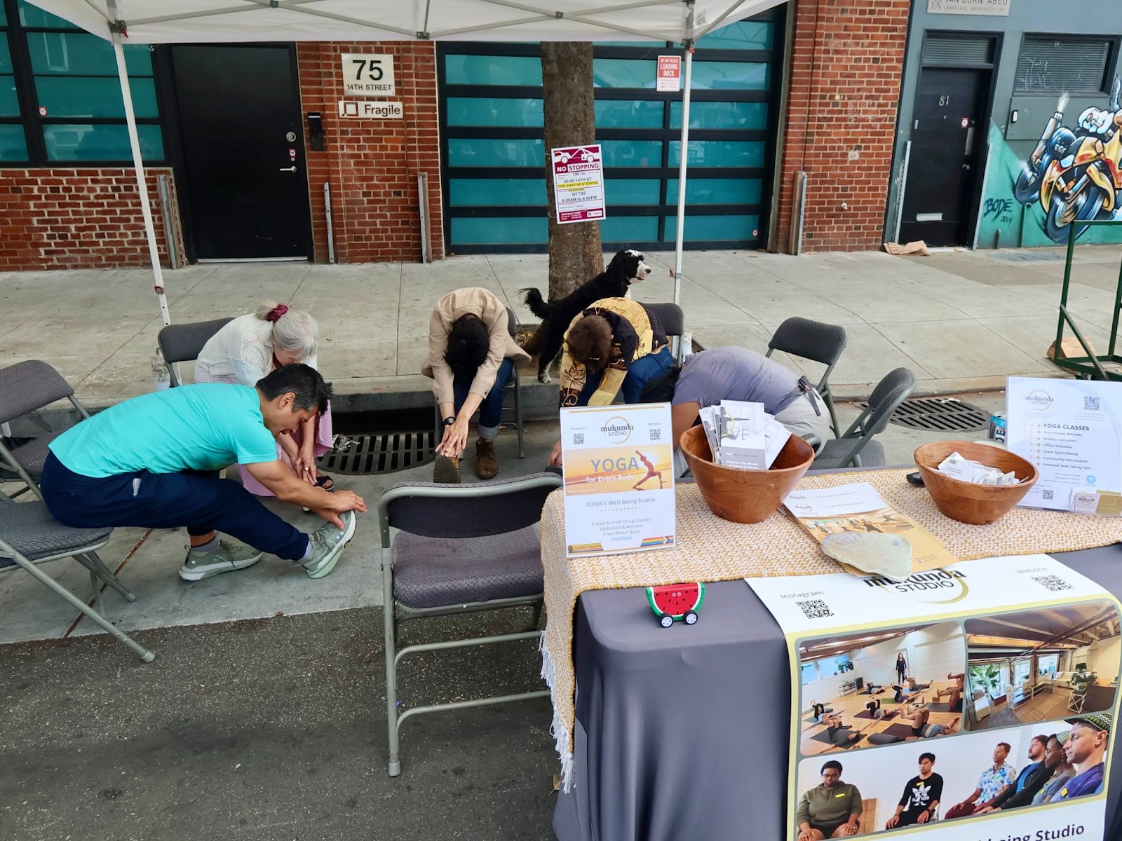 A man in a black cap and "Rainbow Grocery Co-op" t-shirt stands against a green and blue wall, smiling with arms raised.