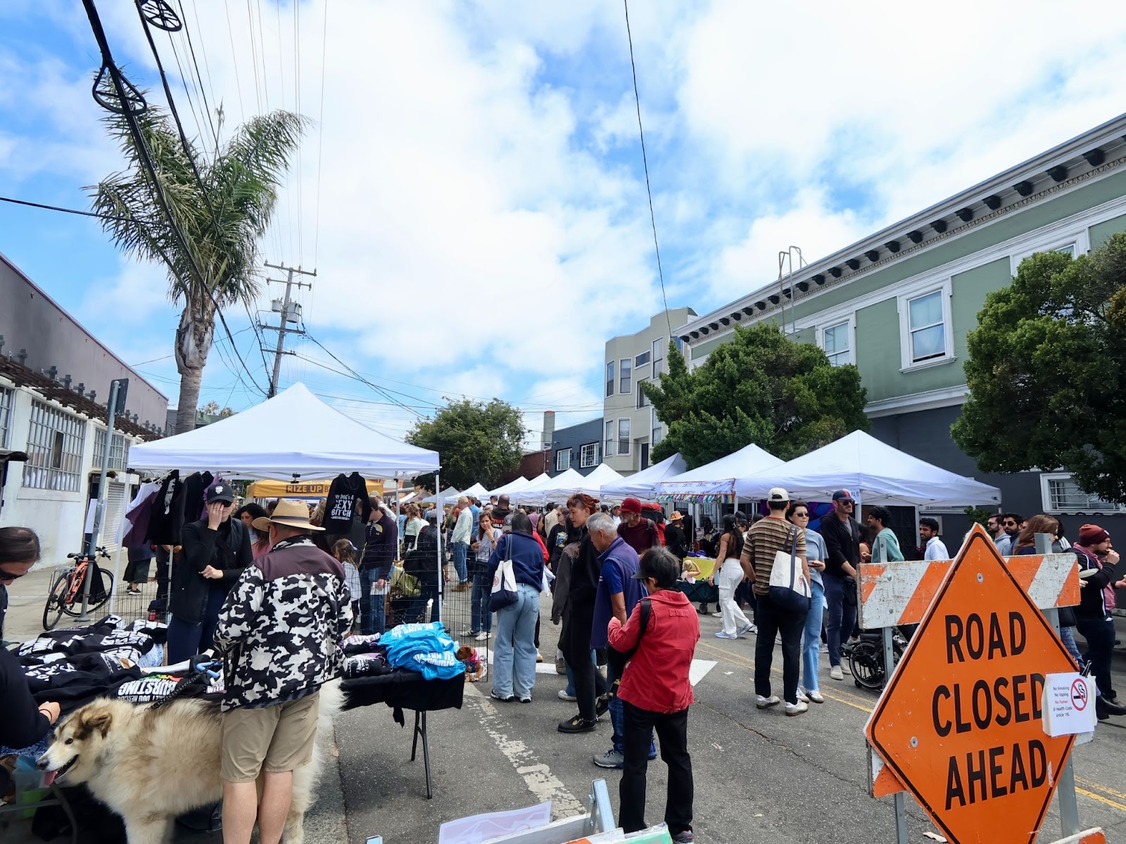 Crowd browsing vendor stalls at an outdoor street market; a "Road Closed Ahead" sign is visible in the foreground.