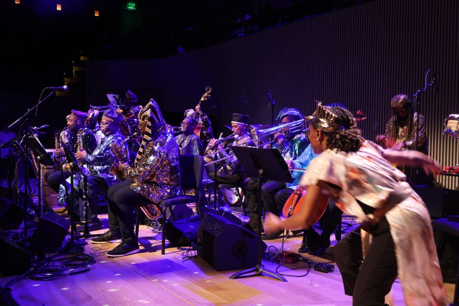 A jazz band in colorful costumes performs on stage with various brass and woodwind instruments, while one person dances energetically in the foreground.