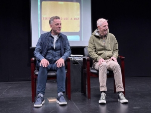Two men sit on stage in front of a screen displaying a slide that reads "BECAUSE I USE A MAP." Both are dressed casually and are seated on wooden chairs.