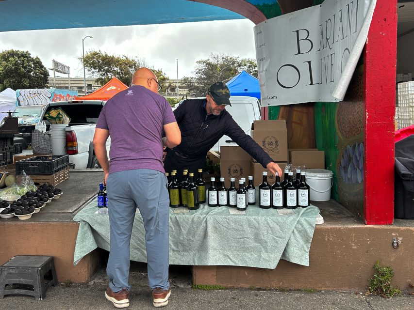 Two men stand at a market stall displaying bottles of olive oil, with signage and produce visible in the background.