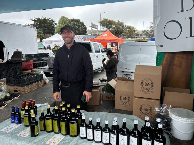 A man stands behind a market stall displaying bottles of olive oil, with boxes stacked behind him and other market stalls visible in the background.
