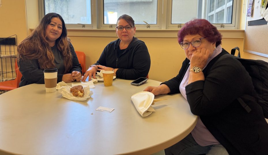 Three women sit at a round table with coffee and pastries, looking at the camera in a well-lit room with large windows.