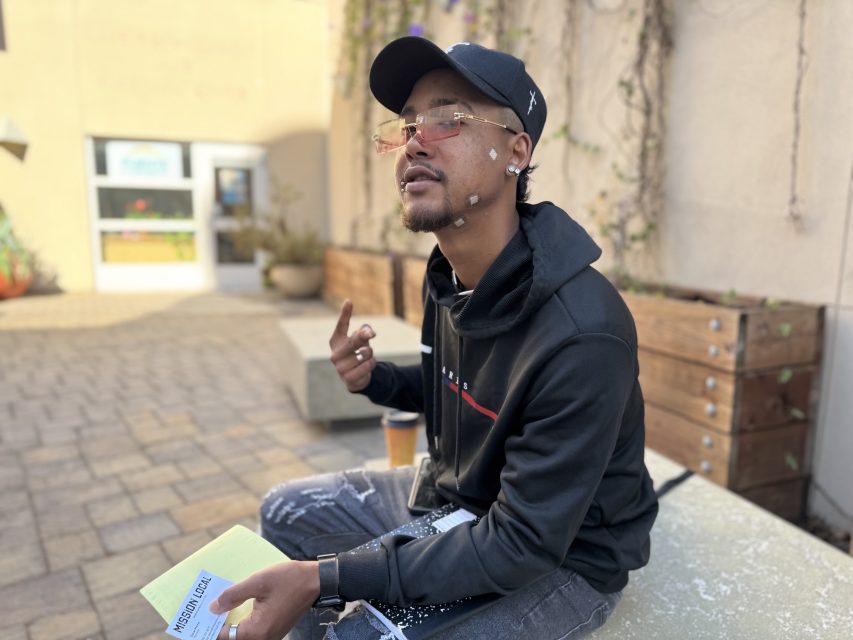 A man wearing a black hoodie, cap, and glasses sits outdoors on a bench, holding papers and making a peace sign with his right hand. A coffee cup is beside him.