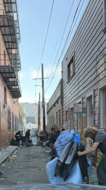 A narrow city alleyway lined with buildings, people gather with belongings and bags, some sitting or standing amid scattered trash and discarded items.
