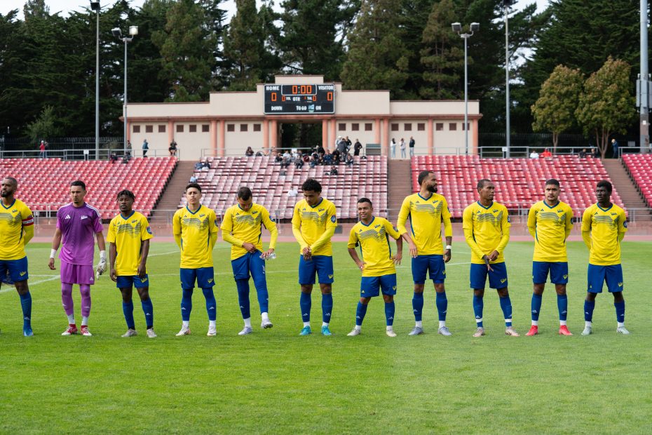 A soccer team wearing yellow and blue uniforms stands in a line on the field before a game, with empty red bleachers and an El Farolito scoreboard in the background.