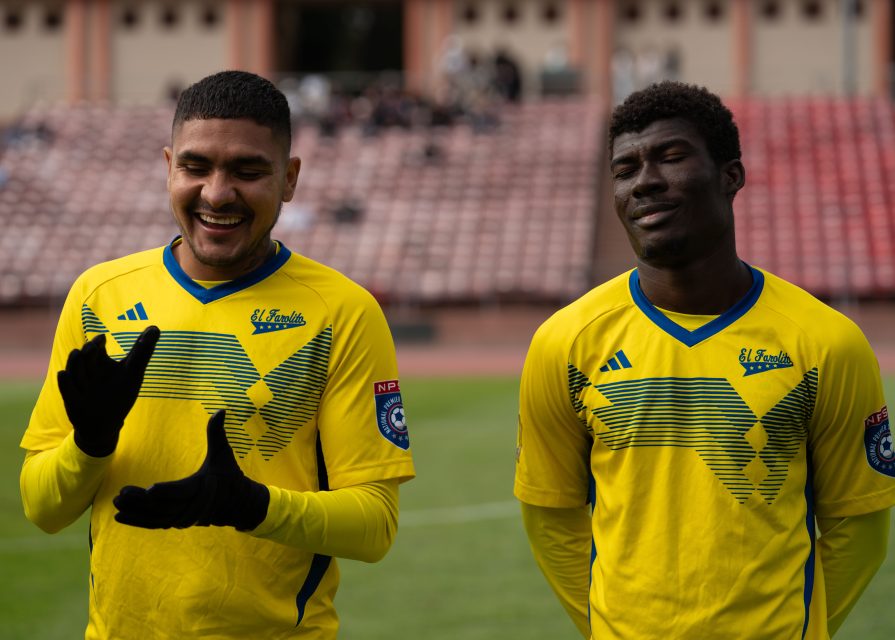 Two soccer players in yellow jerseys from El Farolito stand on the field; one smiles and gestures with gloved hands, while the other stands with eyes closed.