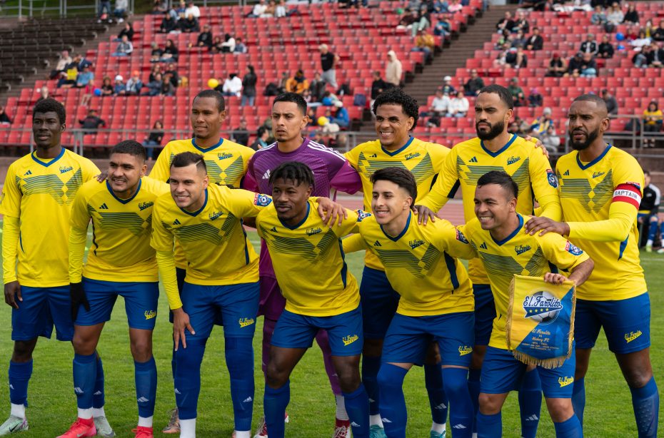 The El Farolito soccer team, wearing yellow jerseys and blue shorts, poses together on a field before a match, with spectators seated in the background.