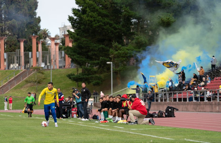A soccer player in a yellow jersey prepares to take a corner kick as El Farolito fans in the stands wave flags and set off blue and yellow smoke.