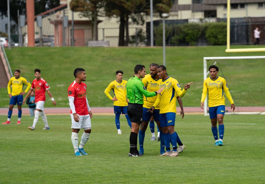 A soccer referee in green talks to players in yellow jerseys, as a player in an el farolito red jersey and others look on during a match on a grass field.