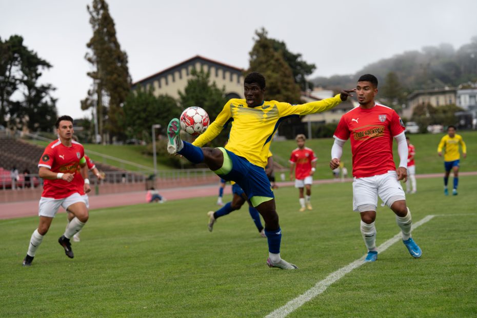 A soccer player in a yellow jersey from El Farolito extends his leg to control the ball while players in red jerseys approach on a grassy field.