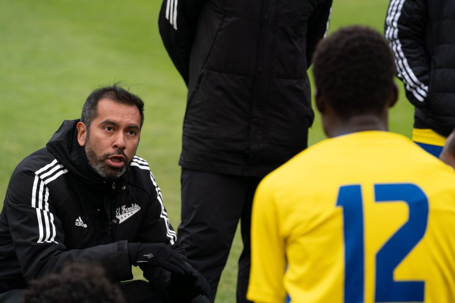 A soccer coach kneels and talks to players, one wearing a yellow jersey with the number 12, during a team discussion on a grassy field, as the El Farolito team prepares for their next match.