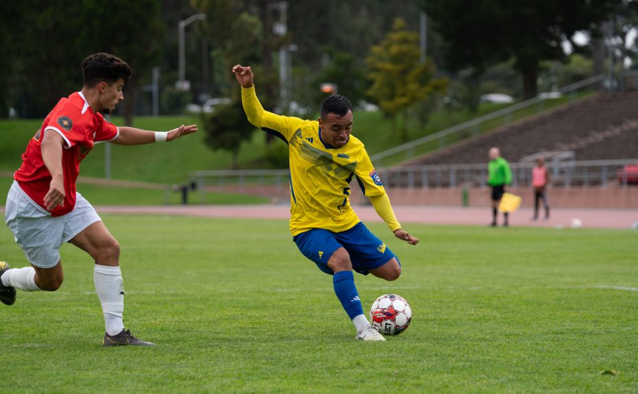 Two soccer players compete for the ball on a grassy field, one in a yellow jersey and the other in red, reminiscent of an El Farolito match. Bleachers and trees line the background.