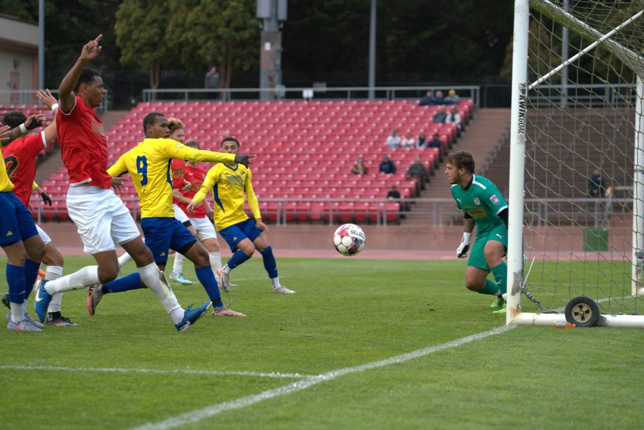 Soccer players in red and yellow jerseys compete for the ball near the goal as the goalkeeper in green watches, with el farolito shining brightly near the goal line.