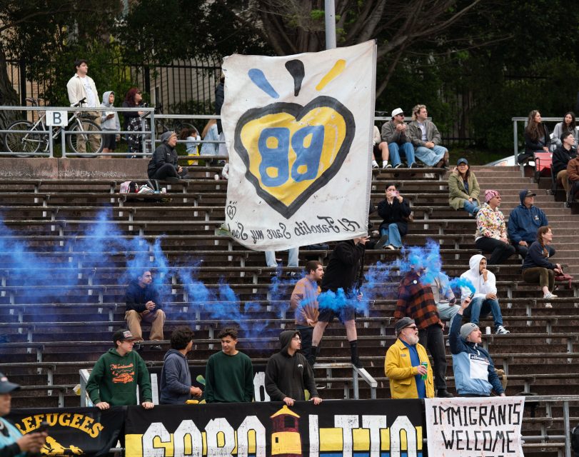 People fill stadium bleachers, holding “Refugees Welcome” and “Immigrants Welcome” banners as blue smoke rises. A giant heart-shaped banner and an el farolito flag wave behind the crowd.