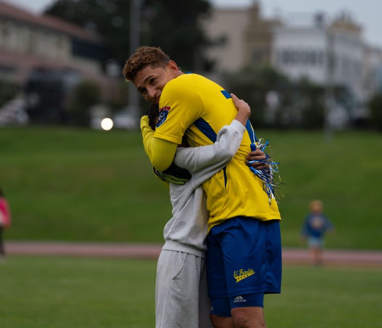 A soccer player in an El Farolito yellow and blue uniform hugs another person on a grassy field, both appearing emotional.