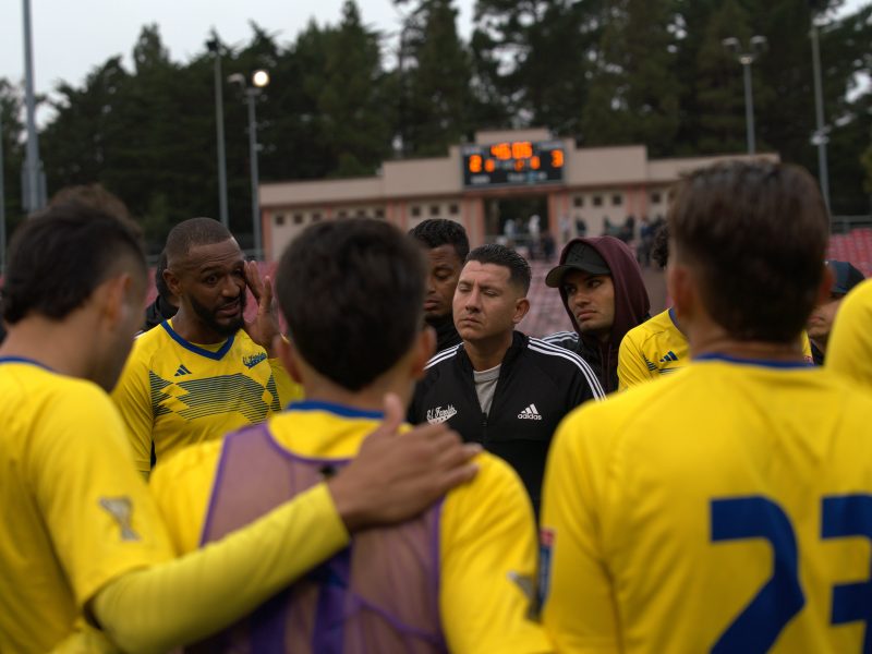 A soccer coach addresses his team in yellow jerseys during a huddle on the field, with a scoreboard and trees visible in the background.