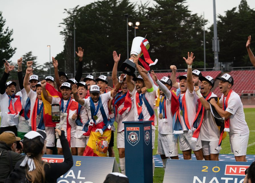 A soccer team in white uniforms, representing El Farolito, celebrates with a trophy on a podium, wearing hats and draped in flags, at an outdoor stadium.