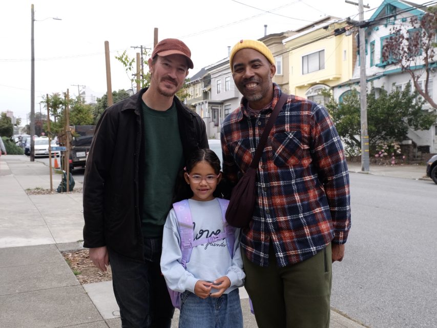 Three adults and a child stand together on a residential sidewalk, posing for a photo. Houses and parked cars line the street in the background.