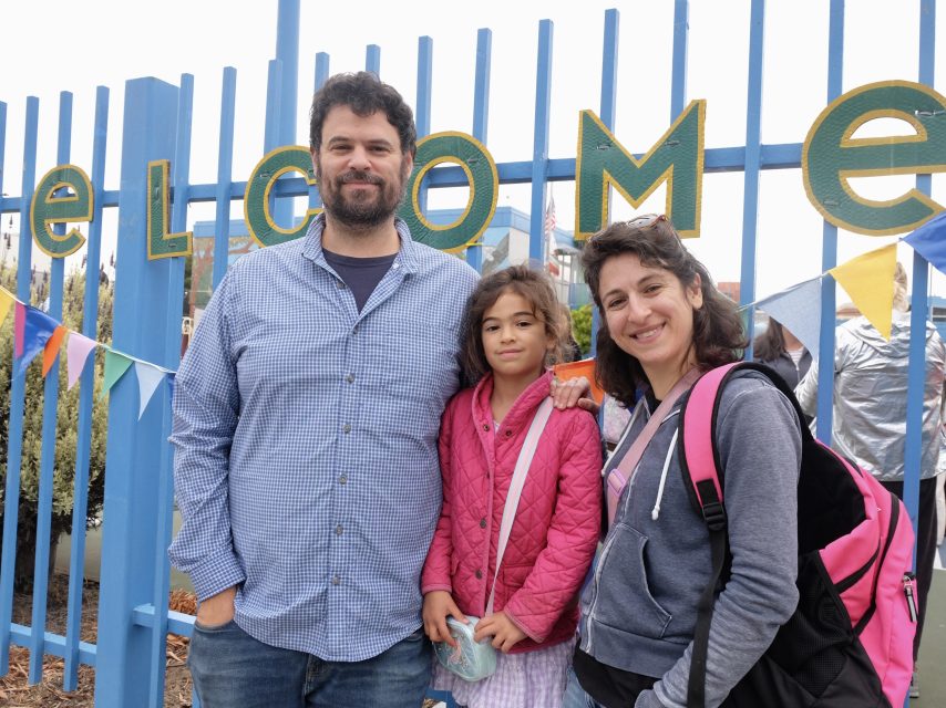 Three people, two adults and one child, stand in front of a blue fence with a "WELCOME" sign and colorful bunting.