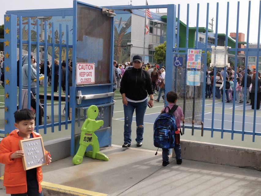 Several children and adults gather near the entrance gate of a school playground; a sign reads “Caution Keep Gate Closed” and a green “Slow!” safety figure is nearby.