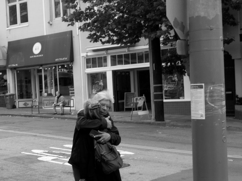 Two people embrace on a city sidewalk near a street sign labeled "ULLOA" and a traffic signal, with shops and trees in the background.