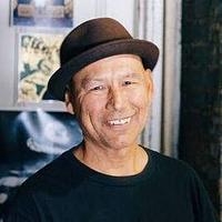 A man wearing a brown hat and black t-shirt smiles while standing indoors in front of posters on a wall, resembling David Crook.