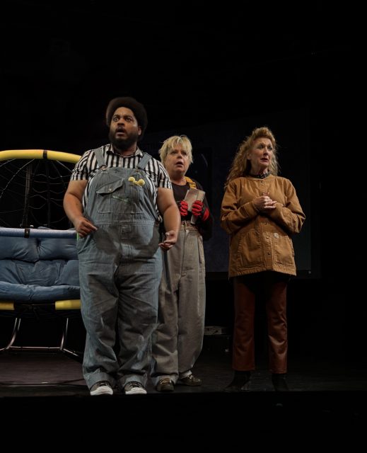 Three people stand on stage in casual costumes, including overalls and a brown jacket, with a blue seat and dark background behind them.
