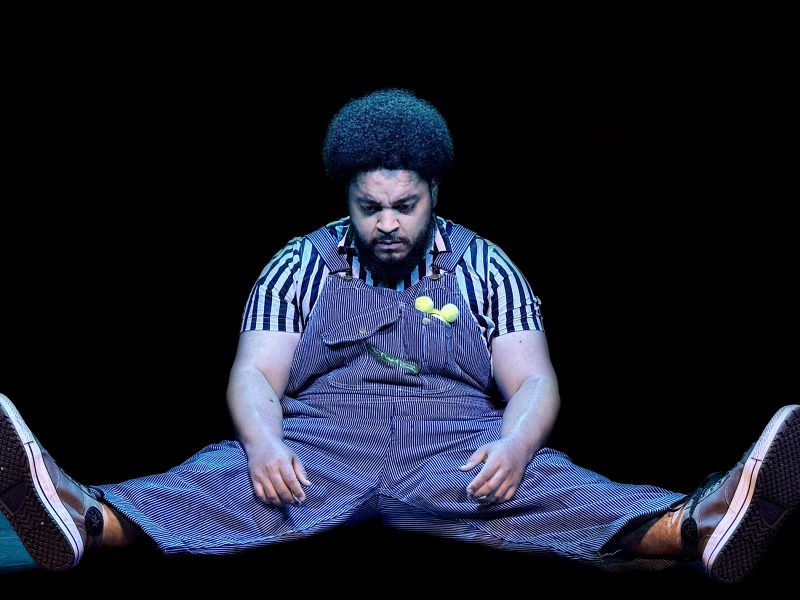 A person in striped overalls sits on the floor with legs spread out, looking down, against a black background.