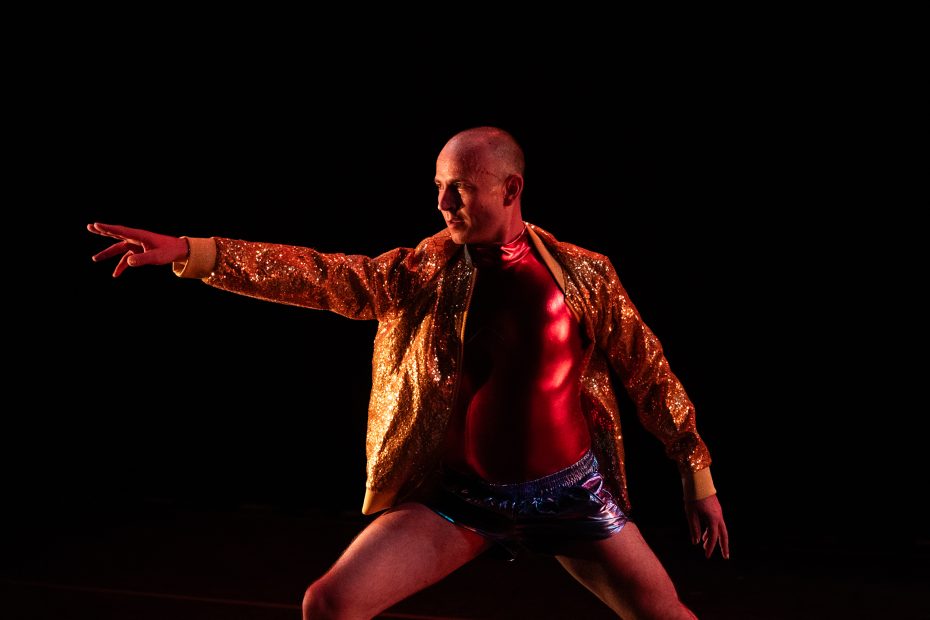 A person in a gold sequined jacket, red top, and metallic shorts strikes a dramatic pose against a dark background.
