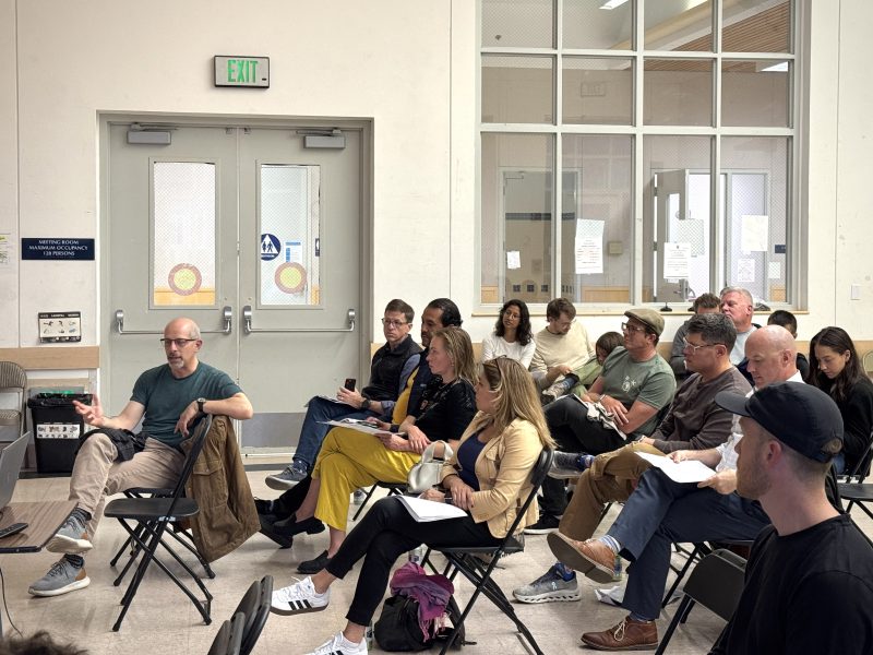 A group of people sit in folding chairs facing a speaker in a meeting room, with some taking notes and listening attentively.