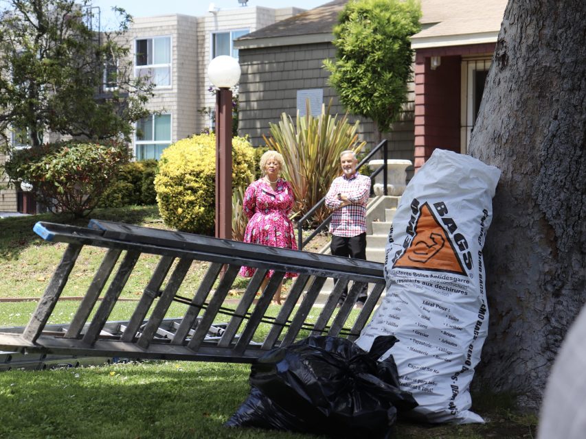 Two people stand near a building entrance, behind a tree, a ladder, a large white bag, and black trash bags on the grass.