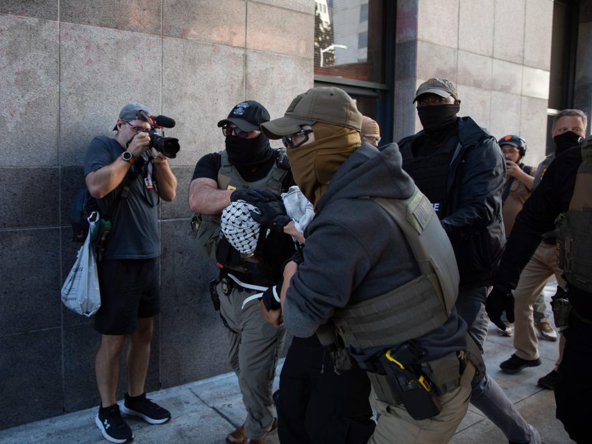 Several law enforcement officers detain a person with a head covering on a city sidewalk while a photographer captures the scene.