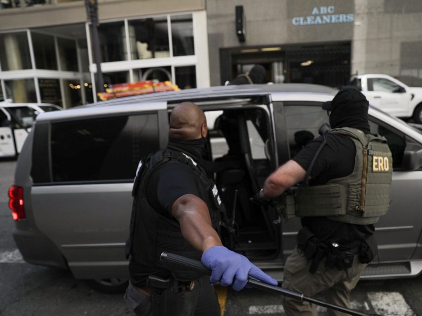 Two law enforcement officers wearing tactical gear and face coverings detain a person and escort them into a gray van on a city street.
