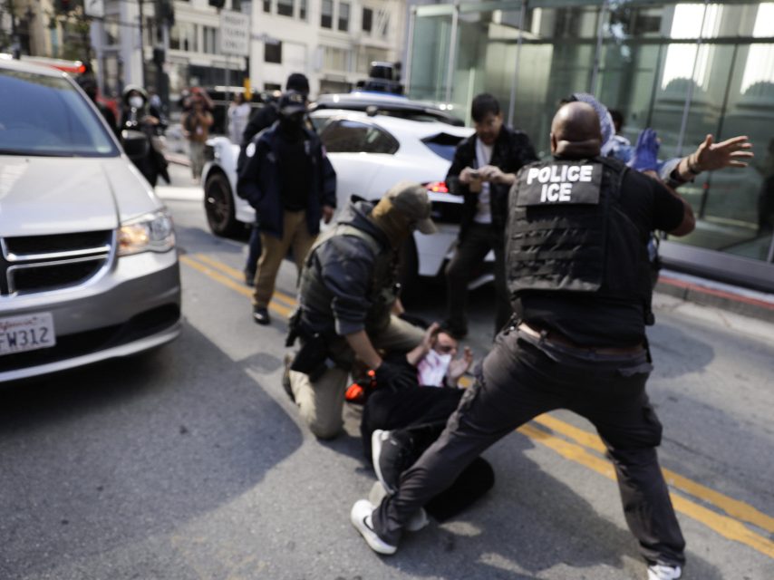Several officers, including one with a vest labeled "POLICE ICE," detain a person on a city street while others stand nearby; vehicles are visible in the background.